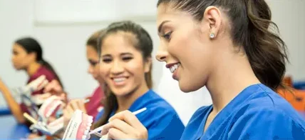 Dental students in blue and maroon scrubs practicing on dental models, smiling and engaged in a classroom setting.