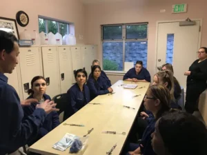 Group of dental students in a classroom setting, seated around a long table with dental instruments, listening to an instructor giving a lesson.