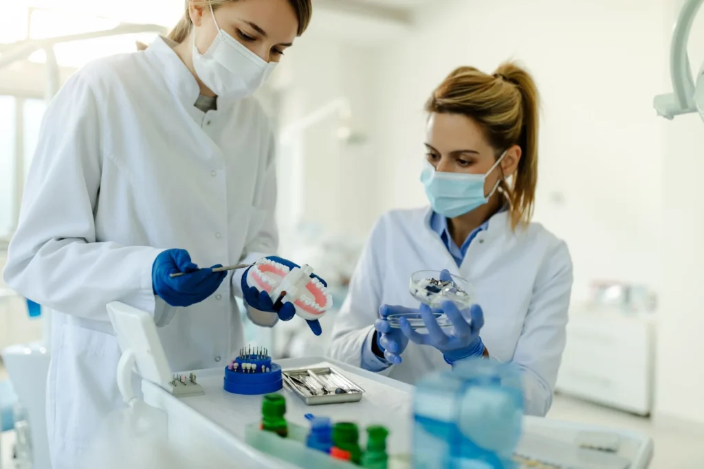 Two dental professionals wearing masks and gloves examining dental tools and a model of teeth in a clinical setting, with various dental instruments on the tray in front of them.