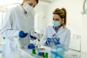 Two dental professionals wearing masks and gloves examining dental tools and a model of teeth in a clinical setting, with various dental instruments on the tray in front of them.