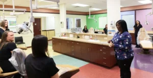 Dental instructor in patterned scrubs teaching a group of students in a clinic setting, demonstrating with a dental model. Students are seated in dental chairs, observing attentively.