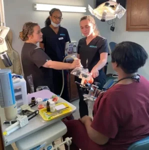 Group of dental students in black scrubs practicing a dental procedure on a model, supervised by an instructor in maroon scrubs.