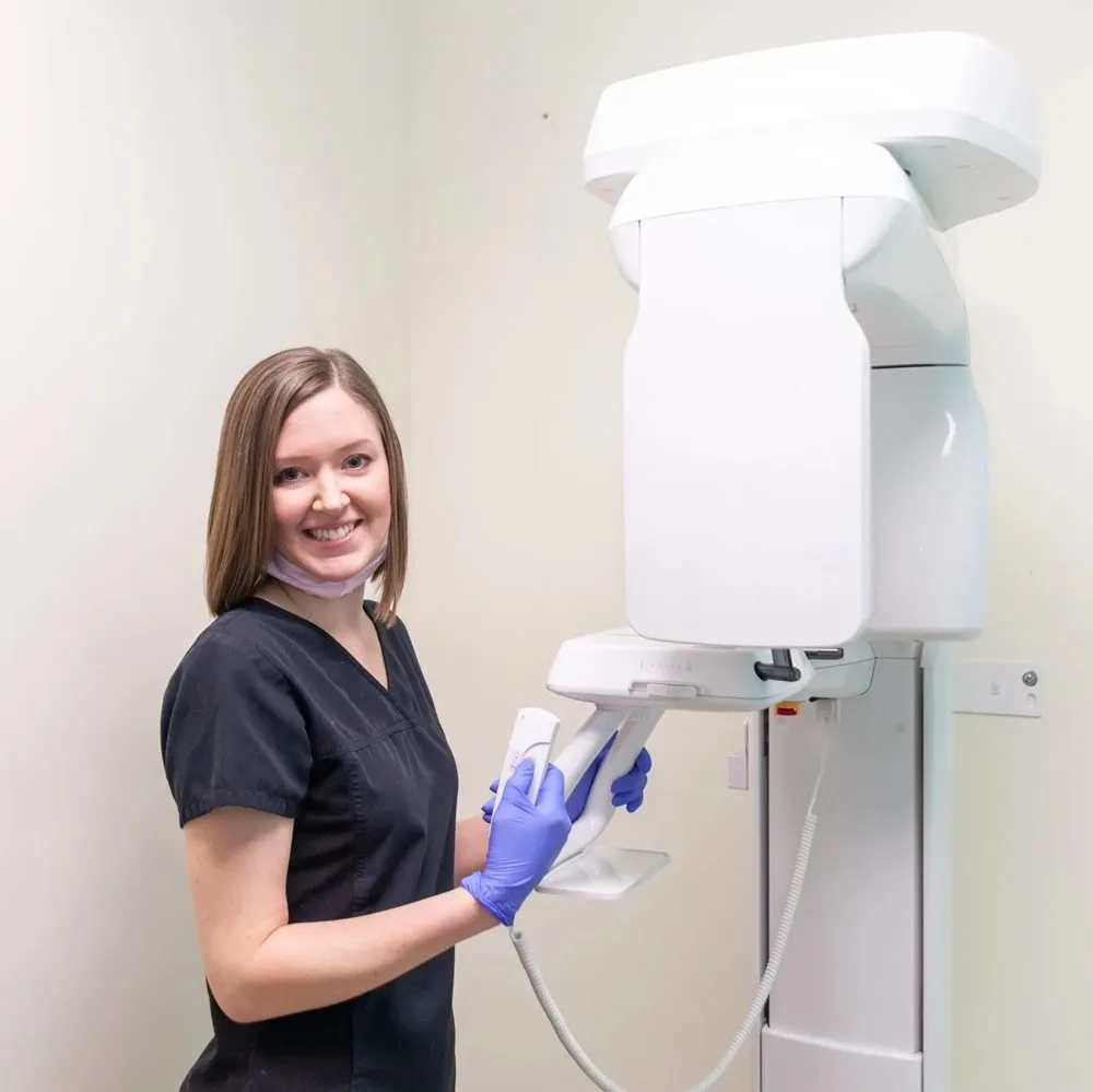 Smiling dental professional in black scrubs and blue gloves preparing a dental X-ray machine.