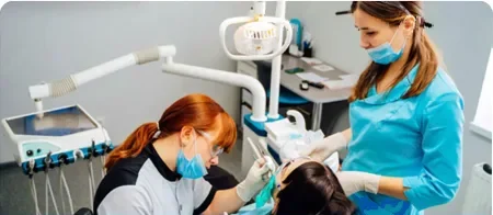 Two dental professionals in masks and gloves working on a patient in a dental chair, with one using dental tools and the other assisting during the procedure.