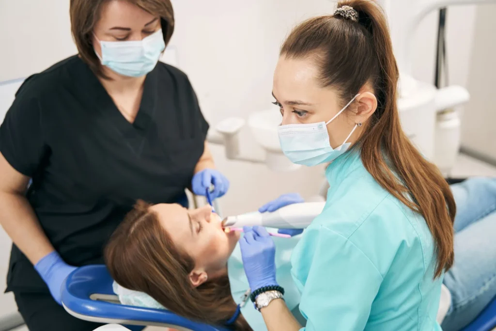 Two dental professionals in masks and gloves performing a procedure on a patient lying in a dental chair. One is holding an intraoral scanner while the other assists.