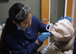 Dental student wearing protective goggles and gloves practicing a dental procedure on a mannequin head in a training environment.