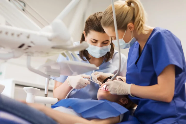 Two dental professionals in scrubs performing a dental procedure on a patient lying in a dental chair, using medical instruments.