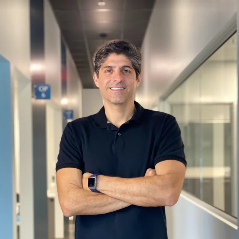 Smiling male professional wearing a black polo shirt and a smartwatch, standing with arms crossed in a modern office hallway.