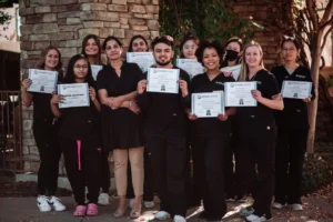 Group of dental assistant students and their instructor posing outdoors, holding certificates of completion, all dressed in black scrubs.