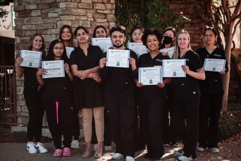Group of dental assistant students and their instructor posing outdoors, holding certificates of completion, all dressed in black scrubs.