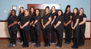 Group photo of a dental office team, all wearing black scrubs, standing in a line inside an office setting. The team members are smiling and posing for the camera.