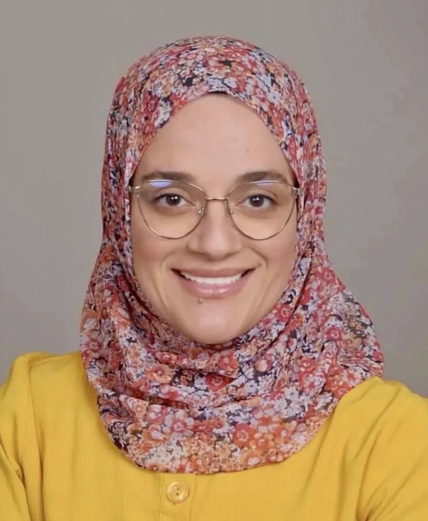 Portrait of Dr. Ingy Aly, a dentist, wearing glasses and a colorful floral hijab with a yellow top, smiling in front of a neutral background.